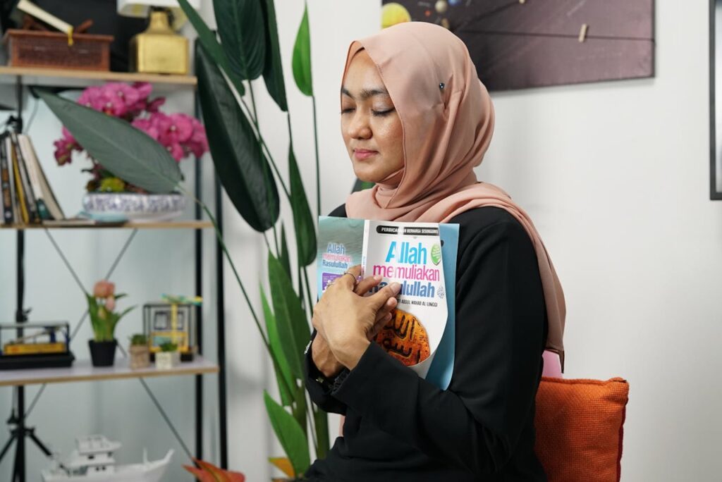 A woman in a hijab peacefully holds a book, eyes closed, in a serene indoor setting.