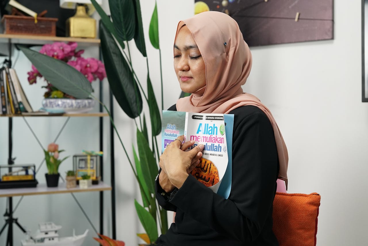 A woman in a hijab peacefully holds a book, eyes closed, in a serene indoor setting.